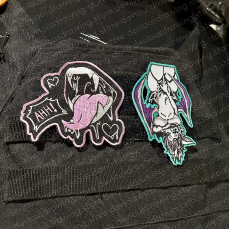 May include: Two embroidered patches on a black fabric surface. The left patch is a black shape with a pink tongue, white teeth, and the text AHH! with two hearts. The right patch is a purple and white winged creature hanging upside down.