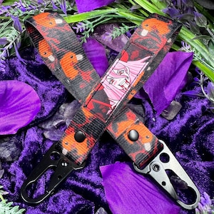 May include: Black keychains with a colorful anime-style design. The keychains feature a black background with orange and red accents, and a pink-haired anime character. The keychains have black metal clasps.