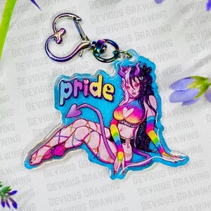Op de afbeelding: Een heldere acryl sleutelhanger met een kleurrijke illustratie van een personage met hoorns, een staart en regenboogaccenten. Het woord "pride" is in een kleurrijk lettertype geschreven. De sleutelhanger heeft een zilverkleurige hartvormige sluiting.