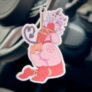 Pode incluir: Um ambientador recortado com um personagem de estilo anime em roupa vermelha e rosa, amarrado com corda. O personagem tem chifres, uma cauda e um pentagrama na parte de trás. O ambientador está segurando um livro intitulado "BDSM GUIDE".