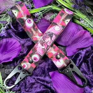 Könnte beinhalten: Zwei schwarze Schlüsselanhänger mit einem rosa-schwarzen Musterband mit Augen und einer Manga-Illustration. Die Schlüsselanhänger haben schwarze Metallverschlüsse und liegen auf einem lila Hintergrund mit Blütenblättern.
