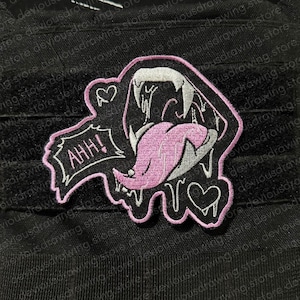 Può includere: Una toppa ricamata raffigurante una bocca aperta stilizzata con zanne bianche affilate e una lunga lingua rosa. Il design include un fumetto con il testo AHH!, due piccoli cuori e un bordo rosa su uno sfondo nero.