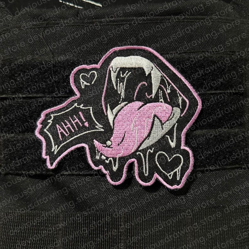 May include: An embroidered patch featuring a stylised open mouth with sharp white fangs and a long pink tongue. The design includes a speech bubble with the text AHH!, two small heart outlines, and a pink border on a black background.