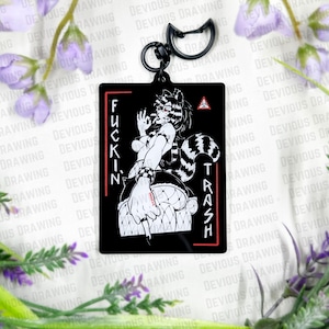 Op de afbeelding: Zwarte rechthoekige sleutelhanger met een gestileerd personage in anime-stijl met een wasbeerstaart. Het personage is in zwart en wit, met de tekst "FUCKIN' TRASH" in een rood kader. Inclusief een zwarte metalen sluiting.