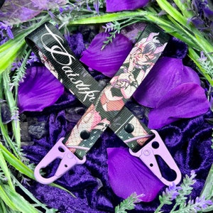 May include: A black lanyard with the name "Satsuki" in white script. The lanyard features a repeating anime-style graphic in pink and white, and is attached to two lavender-colored metal clasps.