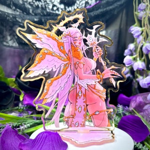 Op de afbeelding: Een roze en gouden acryl standee met een fantasiekarakter met vleugels, een halo en meerdere ogen. Het personage houdt een staf vast. De standee heeft een gouden omtrek en staat op een witte basis met paarse accenten.