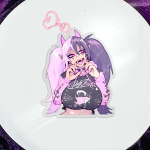 Op de afbeelding: Een heldere acryl bedel met een cartoonfiguur met roze en paars haar, hoorns en hoektanden. Het personage draagt een zwarte top met de tekst "Self Fangs" en een roze jas. Een roze hartvormige sleutelhanger is bevestigd.