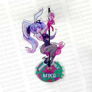 Op de afbeelding: Een heldere acryl figuur met een anime-stijl karakter met konijnenoren, een zwarte outfit en roze accenten. De figuur poseert op een paal, met de tekst "RABBIT HOLE MIKU" op de basis. De achtergrond is wit.