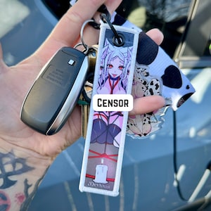 May include: A keychain with a black ring holding a car key, a key tag with a cartoon image, and a cow print charm. The key tag has a white background with a cartoon image of a woman with red eyes and the word "CENSOR" in white.