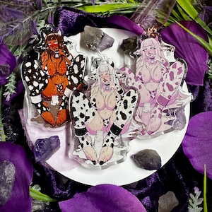 Op de afbeelding: Drie acryl bedels met anime-personages met een koeienthema. Elke bedel toont een personage in een outfit met koeienprint, met een melkfles. De bedels zijn geplaatst tegen een achtergrond van paarse stof, kristallen en groen.