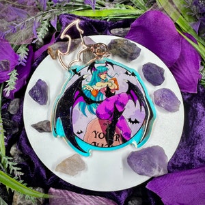 Op de afbeelding: Een ronde, heldere acryl sleutelhanger met een cartoon illustratie van een vrouwelijke vampier met blauw haar, zwarte vleugels en paarse kleding. De sleutelhanger heeft een vleermuisvormige sluiting en de tekst "YOUR GRAVE" op een grafsteen.