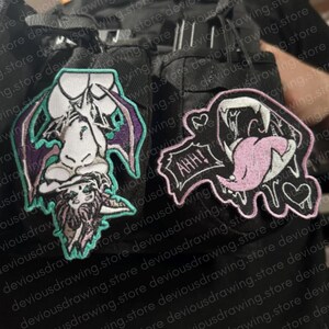 May include: Two embroidered patches attached to a black fabric surface. The left patch depicts a stylised bat-like creature with purple wings and teal outlines. The right patch features a mouth with sharp fangs, a pink tongue, and the text AHH!
