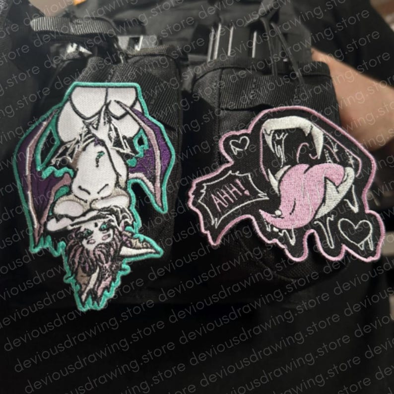 May include: Two embroidered patches attached to a black fabric surface. The left patch depicts a stylised bat-like creature with purple wings and teal outlines. The right patch features a mouth with sharp fangs, a pink tongue, and the text AHH!