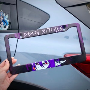 May include: Black license plate frame with purple detailing and the text "DEMON BITCHES" in white. The frame features a purple anime character with red eyes. The frame is held up against a car.