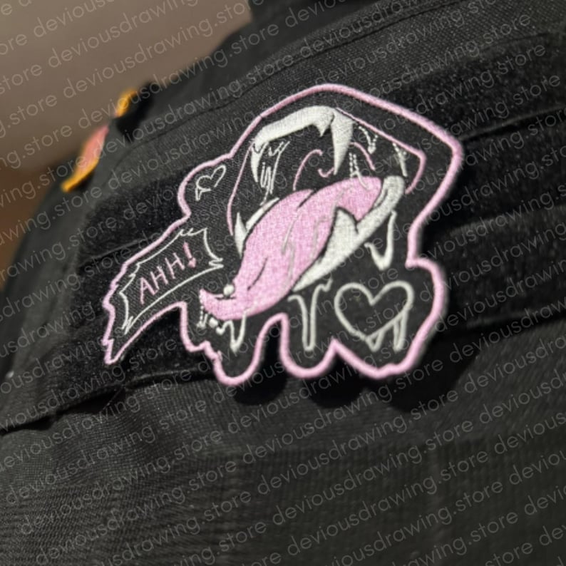 May include: A black embroidered patch featuring a stylised mouth with sharp white teeth and a long pink tongue. The design includes a small heart, a speech bubble with the text AHH!, and a light pink border, all set against a black fabric background.