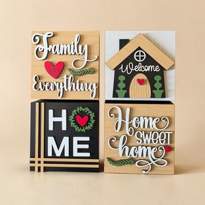 May include: Four decorative wooden blocks with home-themed phrases. One block says "Family is Everything" with a red heart. Another features a house with "Welcome" on it. The third says "HOME" and the last says "Home Sweet Home".