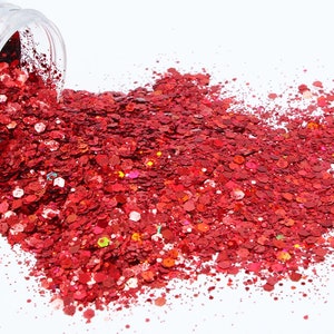 May include: A close-up of a jar of red glitter, with some of the glitter spilling out onto a white surface. The glitter is a mix of red, pink, and gold.