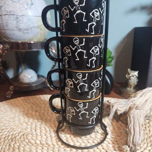 Puede incluir: Un juego de cuatro tazas de cerámica negra apiladas verticalmente en un soporte de alambre metálico negro. Cada taza presenta un estampado gráfico blanco de esqueletos bailando, un borde naranja fino y un asa negra a juego.