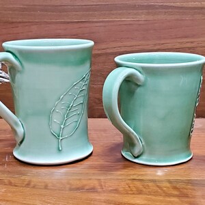 Könnte beinhalten: Zwei handgefertigte, hellgrüne Keramikbecher mit Blattmotiven. Der größere Becher hat einen detaillierten Blatteindruck. Beide Becher haben gebogene Griffe und stehen auf einer Holzoberfläche. Die Becher sind ein passendes Set.