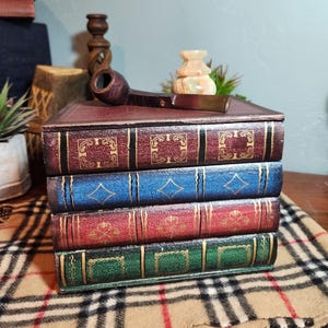 May include: A decorative storage box designed to look like a stack of four vintage books in maroon, blue, red, and green with gold-colored embossed details. A dark brown wooden smoking pipe rests on the top surface of the book-shaped box.