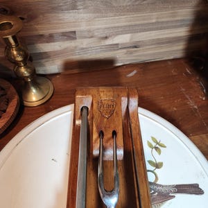 May include: A vintage carving set with a wooden holder and three stainless steel utensils. The holder is engraved with the word "FLINT". The set is displayed on a white tray with a bird and floral design.