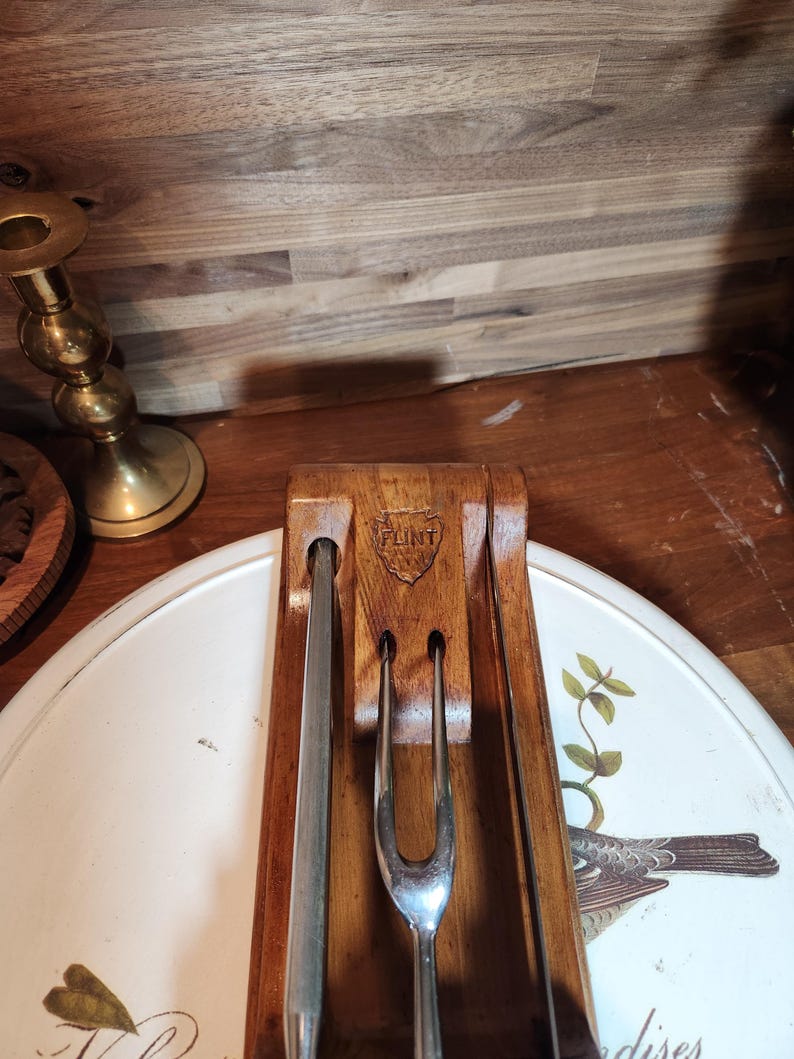 May include: A vintage carving set with a wooden holder and three stainless steel utensils. The holder is engraved with the word "FLINT". The set is displayed on a white tray with a bird and floral design.