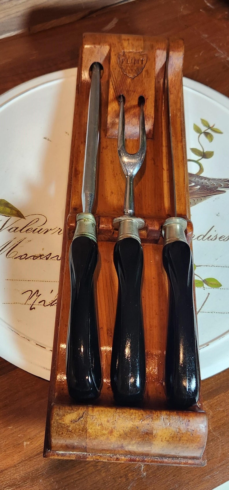 May include: A vintage carving set comprising a carving fork, a carving knife, and a sharpening steel, presented in a wooden holder. The holder is branded with "FLINT". The handles of the utensils are black and the set is for carving.
