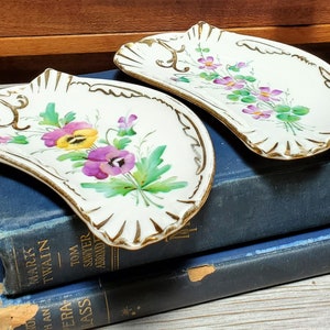Pode incluir: Duas travessas antigas de porcelana em forma de concha com desenhos florais pintados à mão. As travessas apresentam amores-perfeitos roxos e amarelos com folhas verdes, acentuados por detalhes dourados. Estão sobre dois livros azuis vintage.