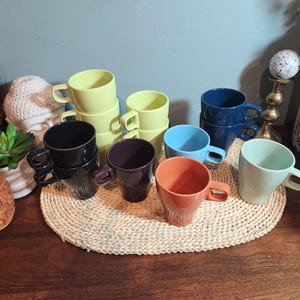 May include: A collection of colorful ceramic mugs in various shades, including yellow, blue, orange, and purple. The mugs are stacked and arranged on a woven mat, suggesting a kitchen or dining setting. The mugs have a simple, modern design.