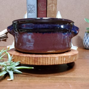 May include: A small, handcrafted ceramic bowl with two handles. The bowl features a deep burgundy base with a dark blue rim. It sits on a round, wooden pedestal, showcasing a rustic aesthetic.