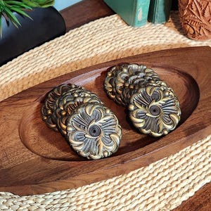 May include: A rectangular wooden tray with a smooth, carved oval center holds two stacks of circular metal drawer knobs. Each knob features a dark, antiqued finish with raised, ornate gold-toned floral patterns and a central screw hole.