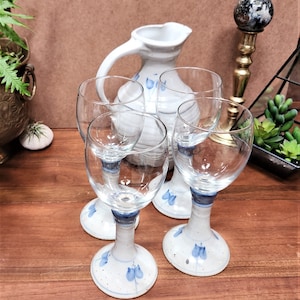 May include: Four clear wine glasses with white ceramic bases and blue floral designs. A matching white ceramic pitcher with a handle and blue floral accents is behind the glasses. The items are on a wooden surface.