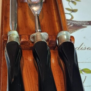 May include: A vintage carving set comprising a knife, a fork, and a carving tool. The utensils feature black handles and silver-coloured metal accents. They are housed in a wooden holder, which has the word "FLINT" engraved on it.
