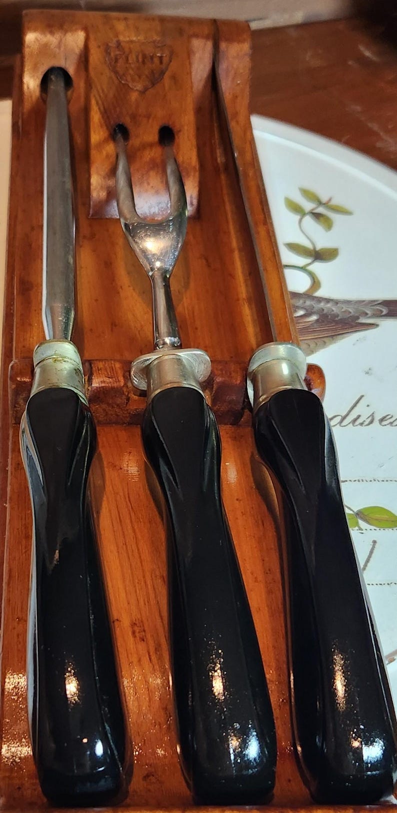 May include: A vintage carving set comprising a knife, a fork, and a carving tool. The utensils feature black handles and silver-coloured metal accents. They are housed in a wooden holder, which has the word "FLINT" engraved on it.