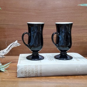 Puede incluir: Dos tazas de vidrio negro con asas y bases de pedestal, cada una con un interior blanco. Las tazas se exhiben sobre un libro antiguo, con un fondo de madera y plantas decorativas. Las tazas son probablemente para café o té.