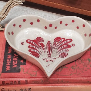 May include: Cream-coloured, heart-shaped ceramic dish with red floral design and polka dots. The dish sits on vintage books. The dish is likely used for serving or display, and is a decorative item.