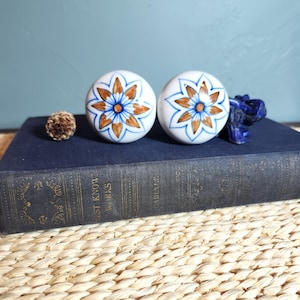 May include: Two white ceramic cabinet knobs with a floral design in blue and gold. The knobs are round and have a glossy finish. A small, brown, textured object and a blue figurine are also present.