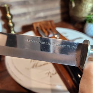 May include: A close-up of a stainless steel knife blade, with the text "FLINT STAINLESS VANADIUM U.S.A." visible. The knife has a shiny, reflective surface and a silver handle. The knife is part of a cutlery set.