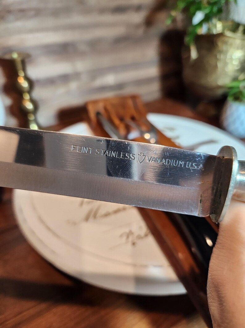 May include: A close-up of a stainless steel knife blade, with the text "FLINT STAINLESS VANADIUM U.S.A." visible. The knife has a shiny, reflective surface and a silver handle. The knife is part of a cutlery set.