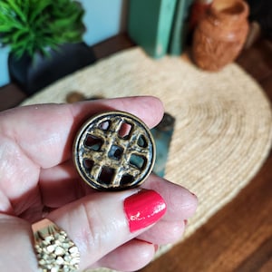 Könnte beinhalten: Ein runder, antiker Messingknopf mit einem geometrischen Gittermuster. Der Knopf wird in einer Hand mit rotem Nagellack und einem goldenen Ring gehalten. Das Design des Knopfes weist sich kreuzende Linien auf, die ein gitterartiges Muster bilden.