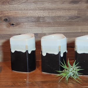 May include: Three square ceramic vases with a black base and a white, dripped glaze. The vases are arranged on a wooden surface with a white Buddha head statue and a small plant in the background. The vases are decorative.