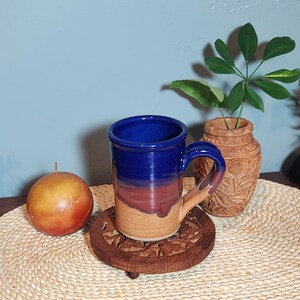 May include: A handmade ceramic mug with a blue top, transitioning to red and tan. The mug sits on a carved wooden coaster. A candle and a decorative vase with a plant are also present.