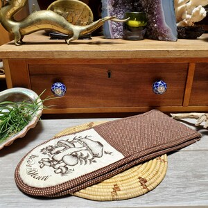May include: A brown and beige oven mitt with a mushroom design and the text "Red Devil". The mitt is on a woven trivet. A wooden drawer with blue and white knobs is in the background.