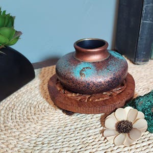 May include: A small, round ceramic vase with a metallic copper and turquoise patina finish. The vase features a stylized Kokopelli figure etched into the side. It rests on a circular, carved wooden base with a dark brown, polished finish.