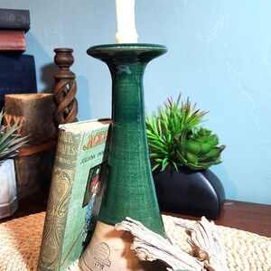 May include: A tall, conical ceramic candle holder with a glossy dark green glaze on the upper portion and a matte, unglazed tan base. The base features a stamped logo and the year 1993. A white taper candle is inserted into the top opening.