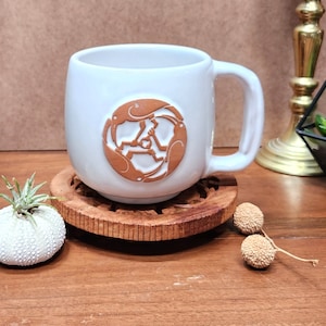 May include: A white ceramic mug with a brown circular emblem featuring a stylized design. The mug sits on a carved wooden coaster. Decorative elements include a small plant in a white shell and dried seed pods.
