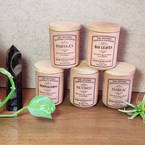 May include: A collection of six pink ceramic canisters with wooden lids, labeled with the names of various spices. The text on the canisters reads "The Revised Suffolk Canister" and the names of the spices. The canisters are arranged on a wooden surface.