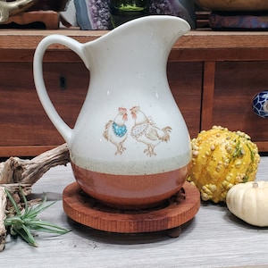 May include: Cream-colored ceramic pitcher with a brown base and a handle. The pitcher features a hand-painted design of two chickens wearing decorative accessories. The pitcher sits on a wooden coaster, with a small gourd and pumpkin nearby.