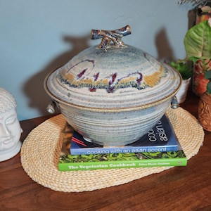 May include: A large, round ceramic serving dish with a lid. The dish is blue and gray with brown and purple accents. The lid has a decorative handle. The dish sits on a woven placemat with two cookbooks underneath.