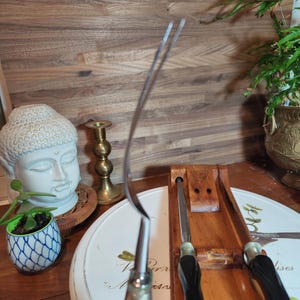 May include: A stainless steel fork with a curved design, held in front of a wooden knife block with two knives. The fork has a black handle. A white Buddha statue and a small potted plant are in the background.
